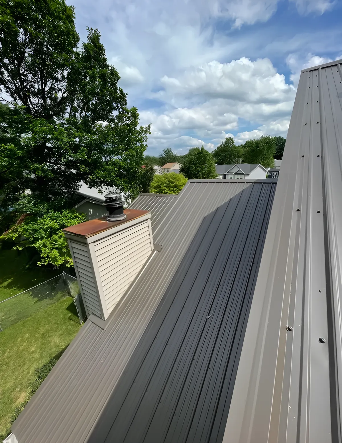 Rooftop with gray metal panels beside a brick chimney, surrounded by trees and a cloudy sky.