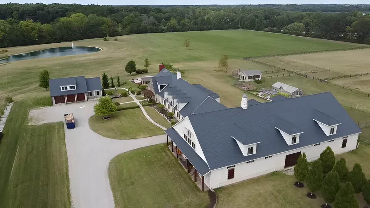 Aerial view of a large countryside house with a gray roof, driveway, lawn, and surrounding fields and trees.