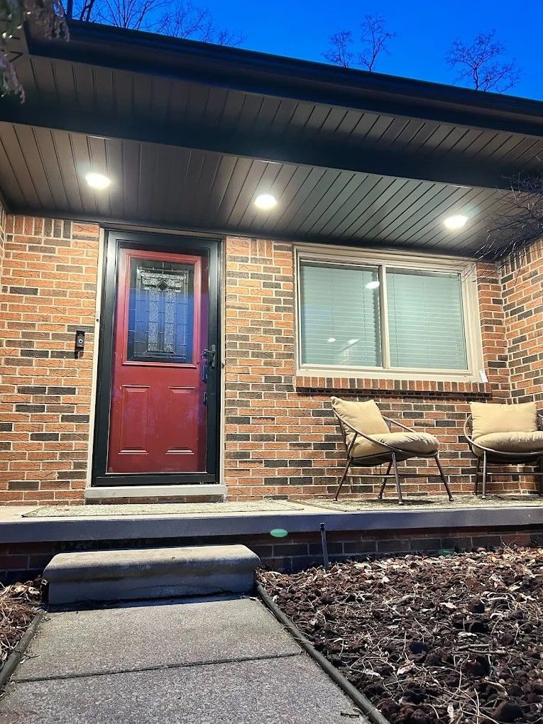 Front porch of a brick house at dusk with a red door, lit ceiling lights, and two chairs by a window