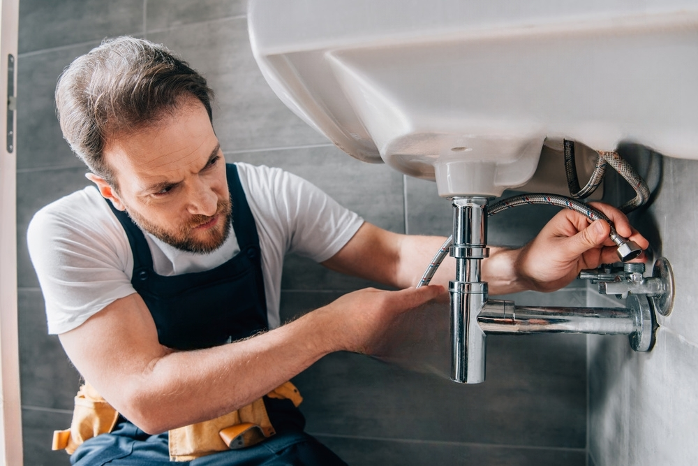 Plumber working under a white bathroom sink, connecting pipes, in a bathroom with gray tiles.