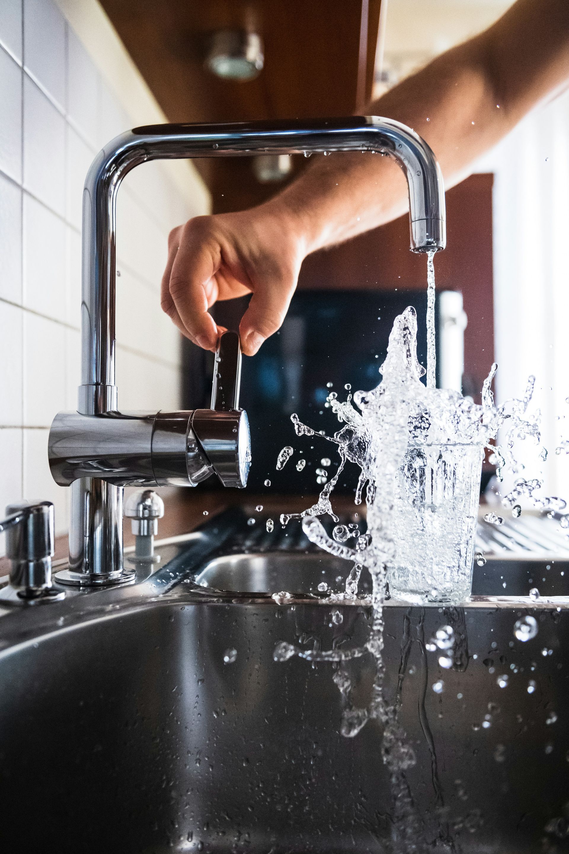Hand turning faucet, splashing water into a glass in a kitchen sink.