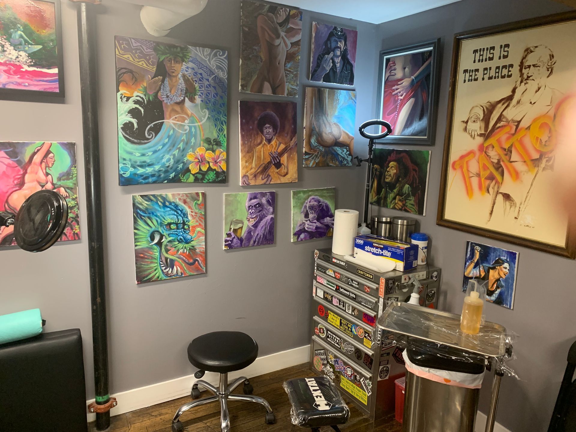 Tattoo studio interior. Artwork covers gray walls. Black chair, stainless steel workstation, and art supplies.