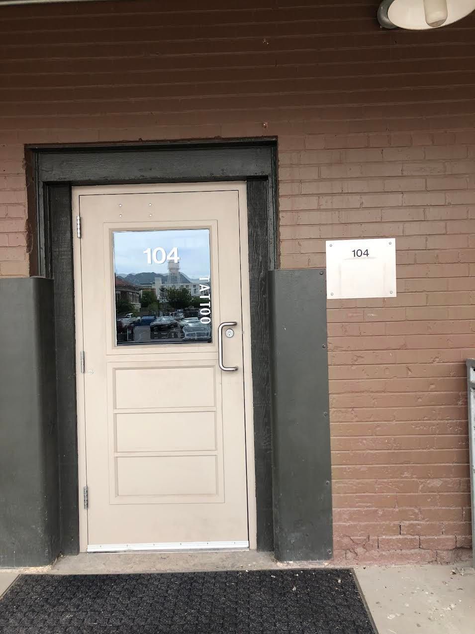 Exterior door, building number 104, tan door with window, brick wall, outdoor setting.