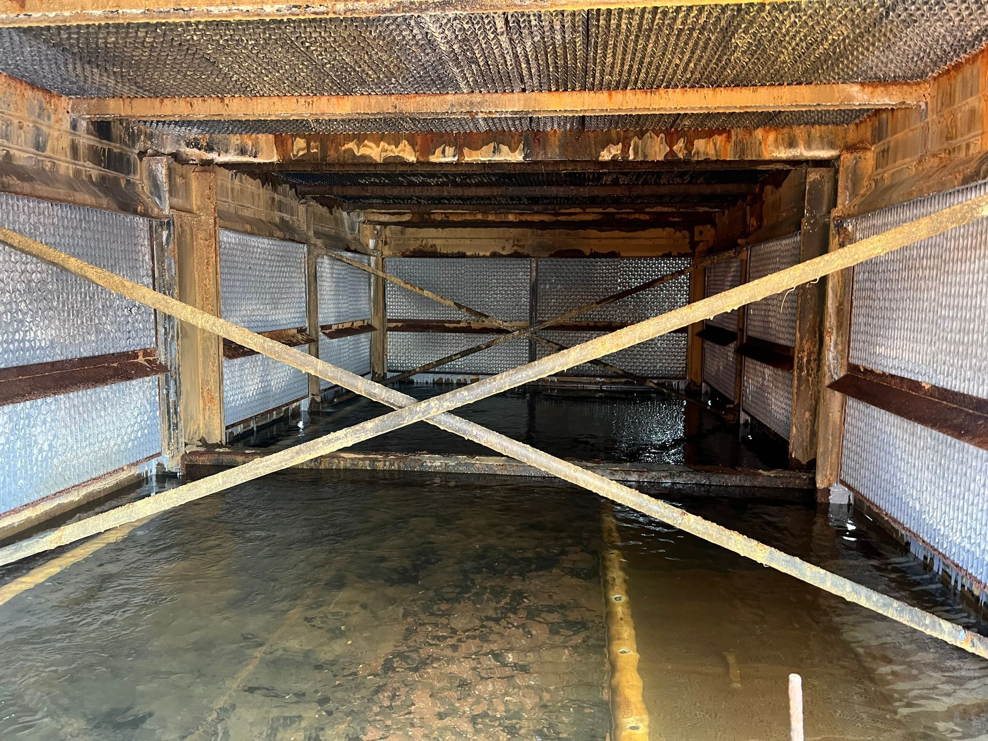 Inside a large mesh enclosure, wooden cross-bracing spans over a shallow water channel.