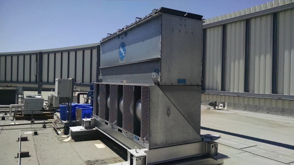 Large industrial cooling unit on a rooftop, with fans and a corrugated metal backdrop under a sunny sky.