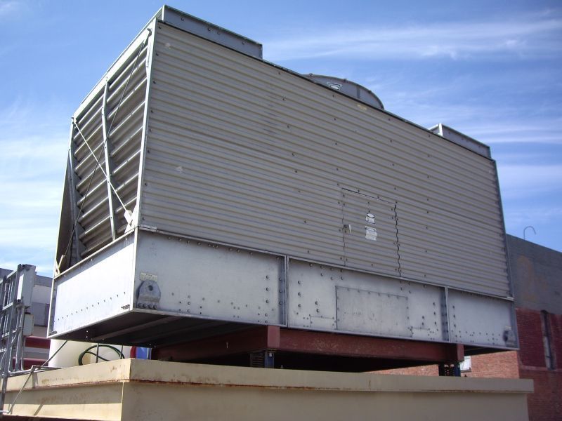 Cooling tower on a rooftop against a blue sky. Gray metal with louvers, mounted on a tan base.