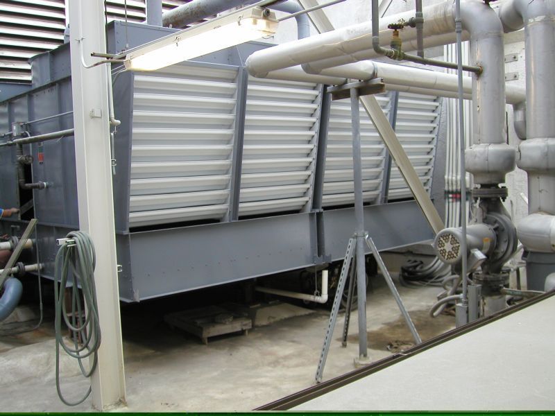 Large industrial cooling tower, grey, with angled louvers, pipes, and metal support frame.