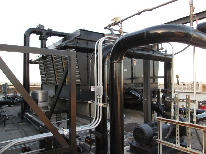 Cooling tower on a rooftop, with black pipes, electrical conduit, and metal grating.