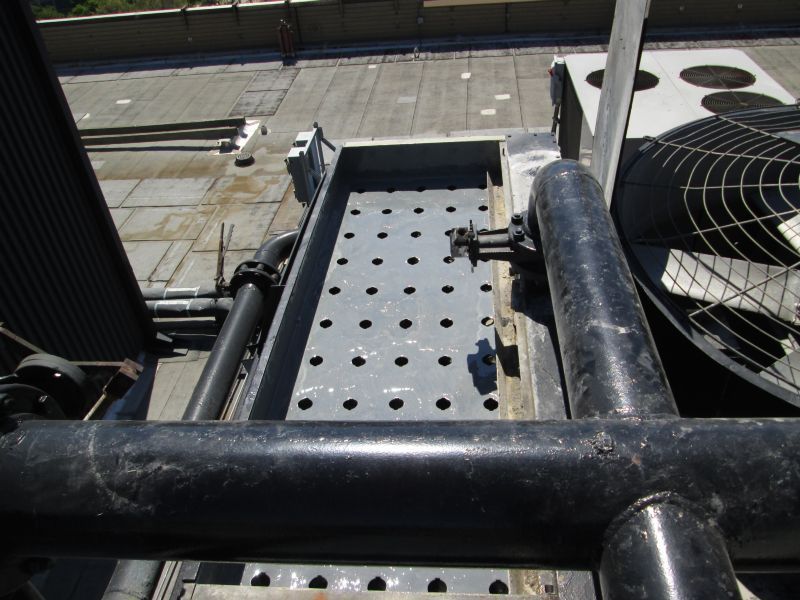 Overhead view of HVAC equipment on a rooftop: black pipes, metal grating, cooling fan, and a gray rooftop.