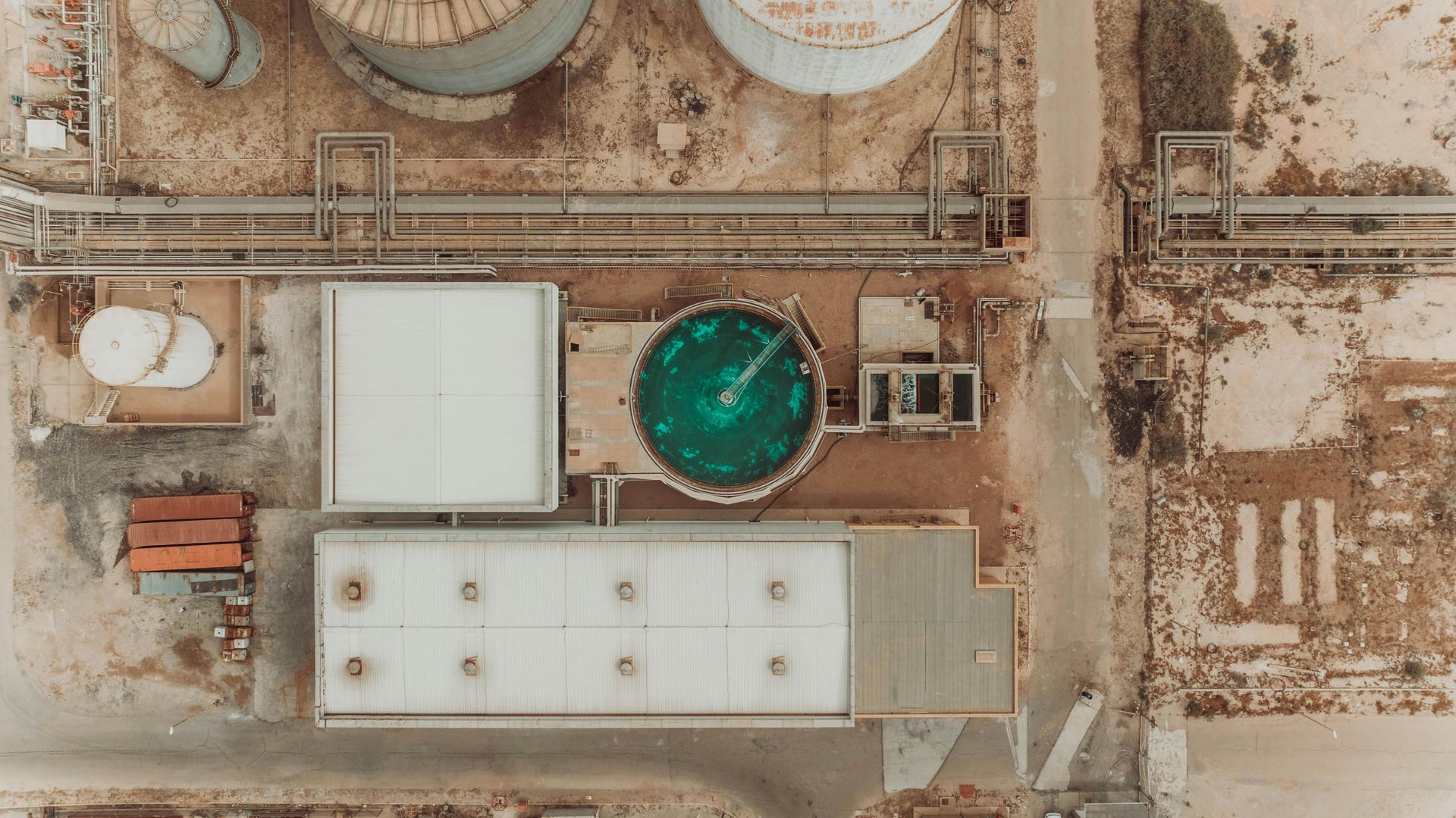 Overhead view of an industrial facility with tanks, buildings, and a large circular green pool.