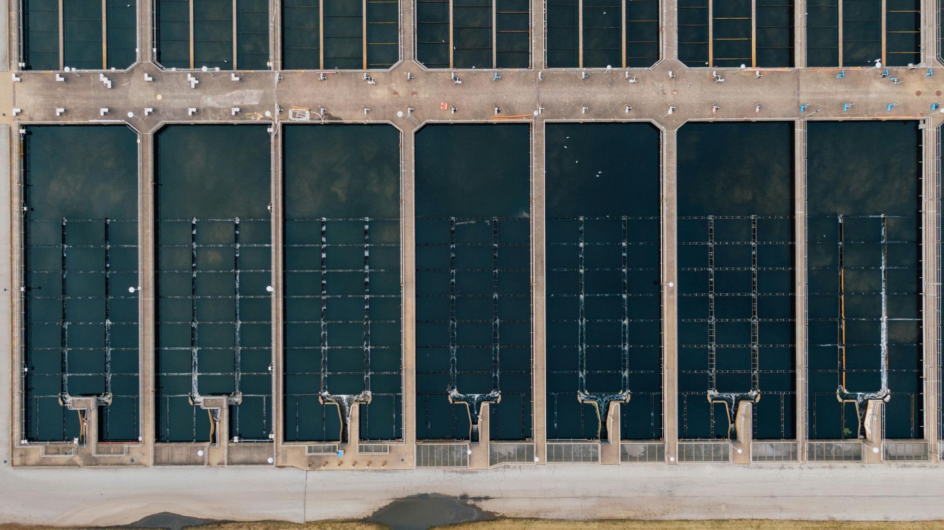 Aerial view of a wastewater treatment plant with rectangular settling tanks, concrete structures, and dark water.