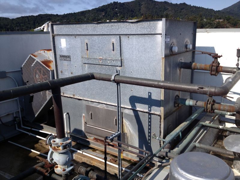 Rooftop HVAC unit with metal casing, pipes, and fan, against a backdrop of mountains and sky.