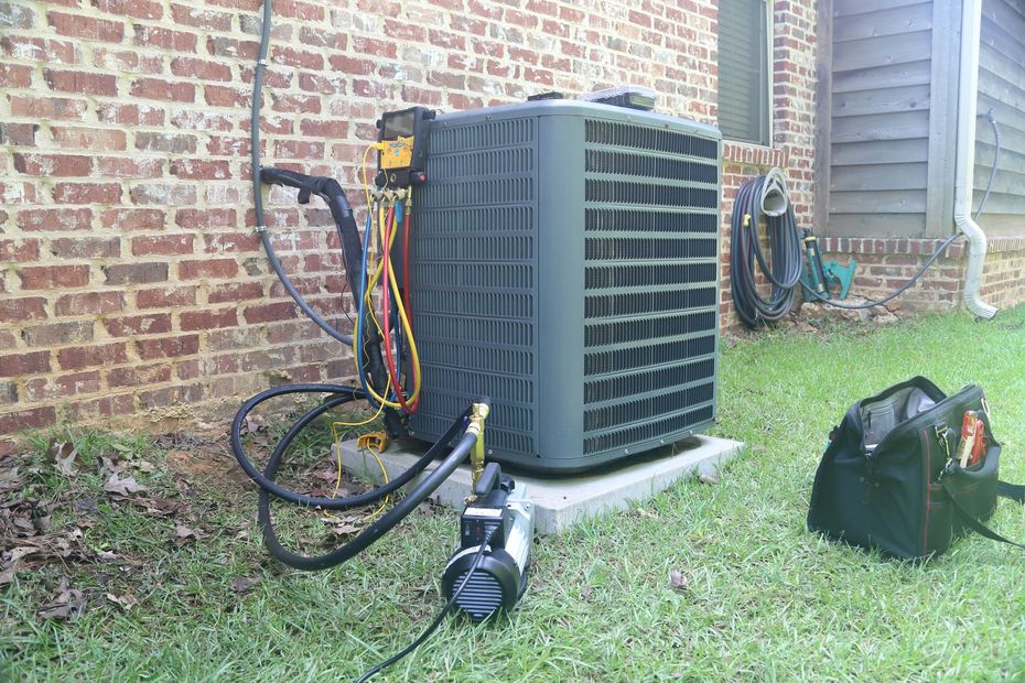 Air conditioning unit being serviced outdoors, near a brick wall and a house.