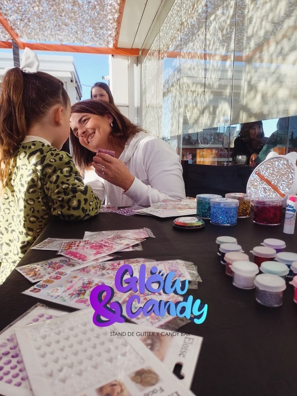 Puesto de pintacaras con un niño al que una mujer le está pintando la cara, materiales coloridos sobre la mesa.