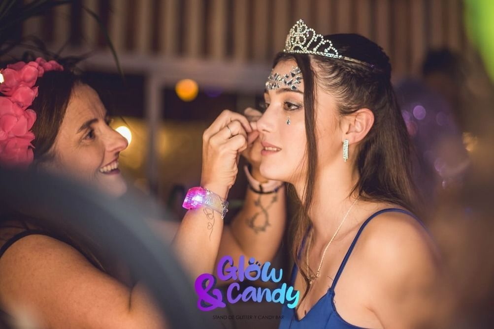 Mujer maquillando a otra mujer que lleva una tiara en una fiesta, con luces borrosas de fondo.