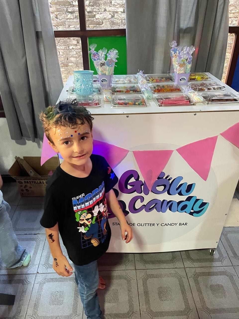 Un niño está de pie junto a una mesa de dulces con golosinas de colores pastel y una pancarta que dice 