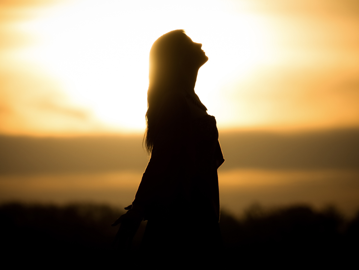 A silhouette of a person standing with their head tilted back against a bright, warm sunset.