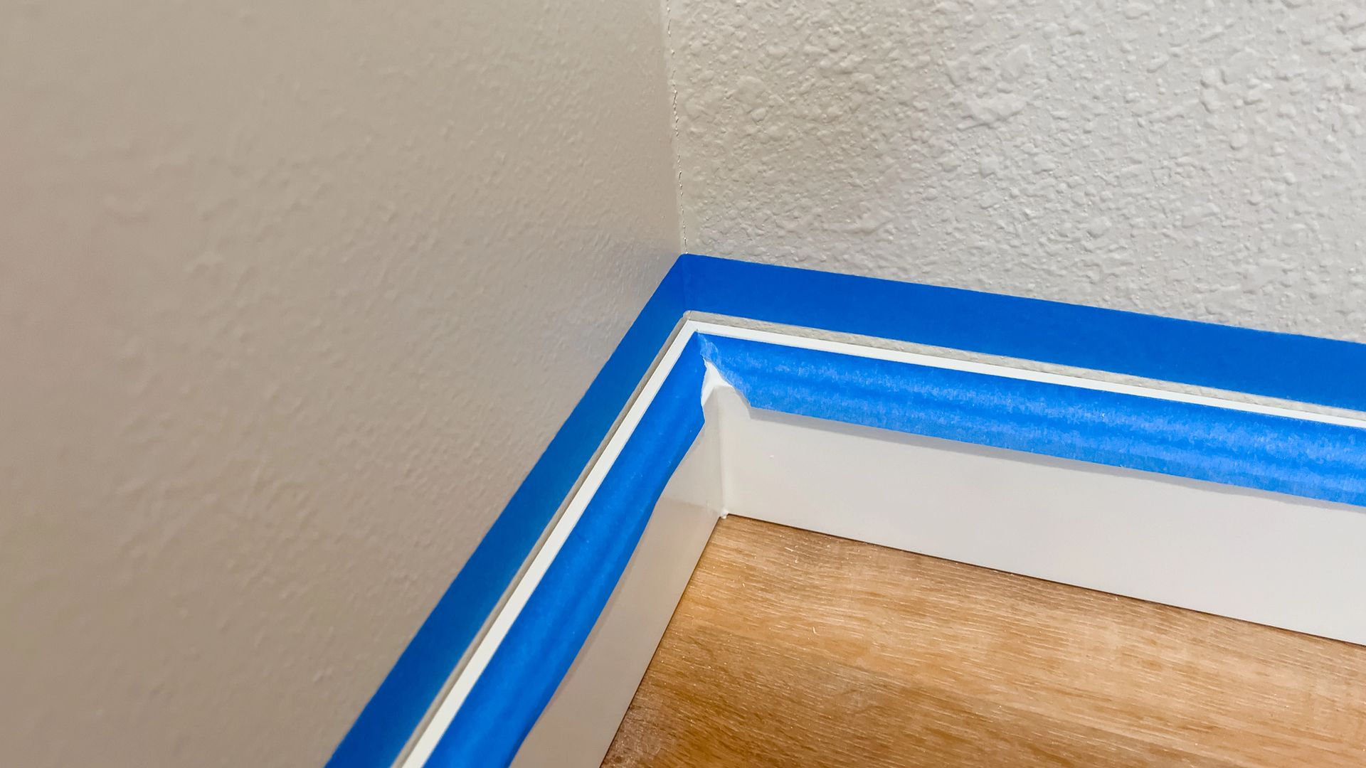 Blue painter's tape lines a baseboard corner on a wood floor, preparing for painting.