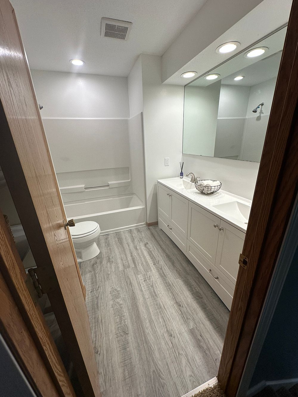 Bathroom with white cabinets, large mirror, tub, and gray wood-look flooring.