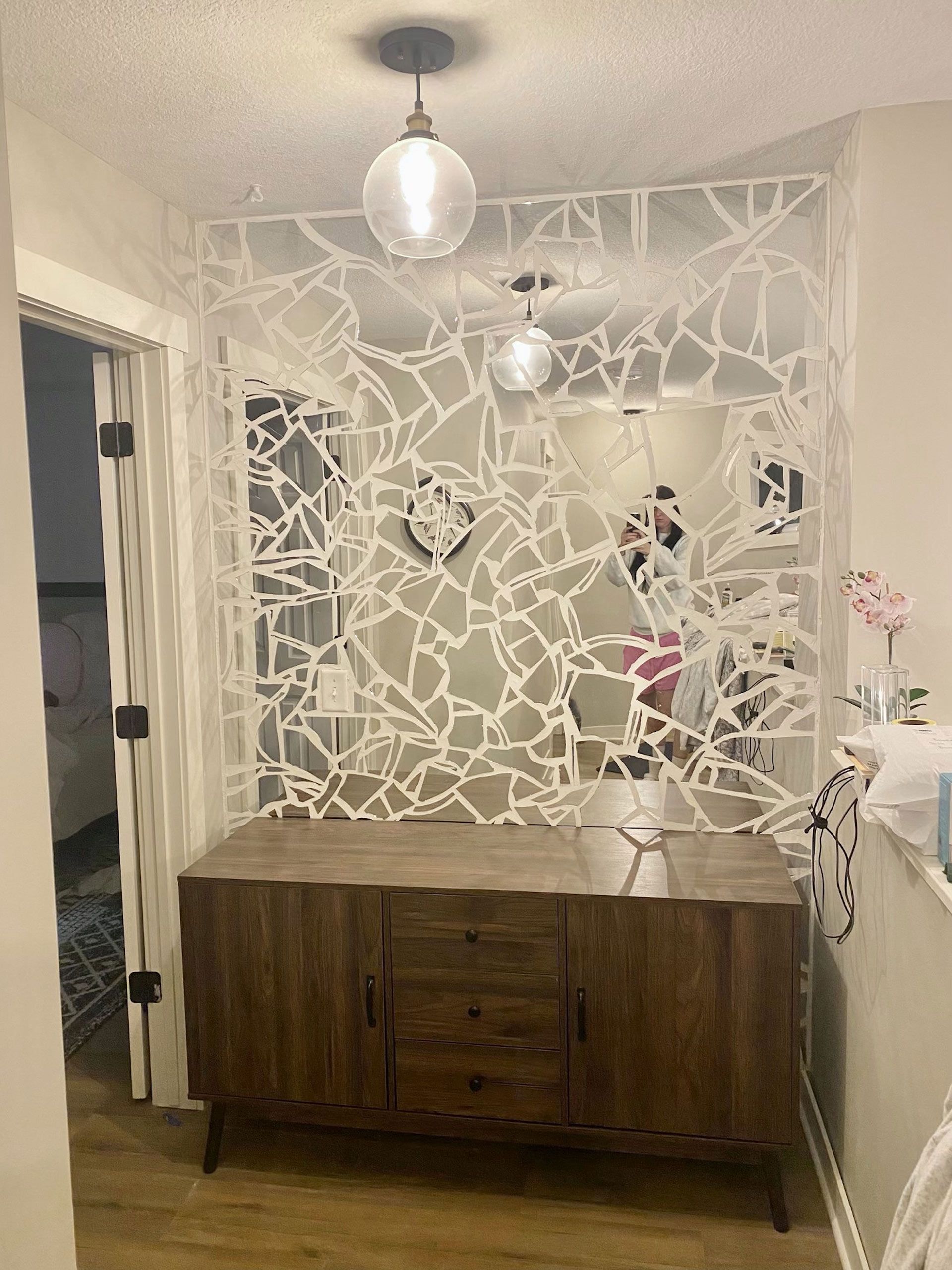 A wooden console table with drawers beneath a decorative white wall with a fractured design. A light fixture hangs above.
