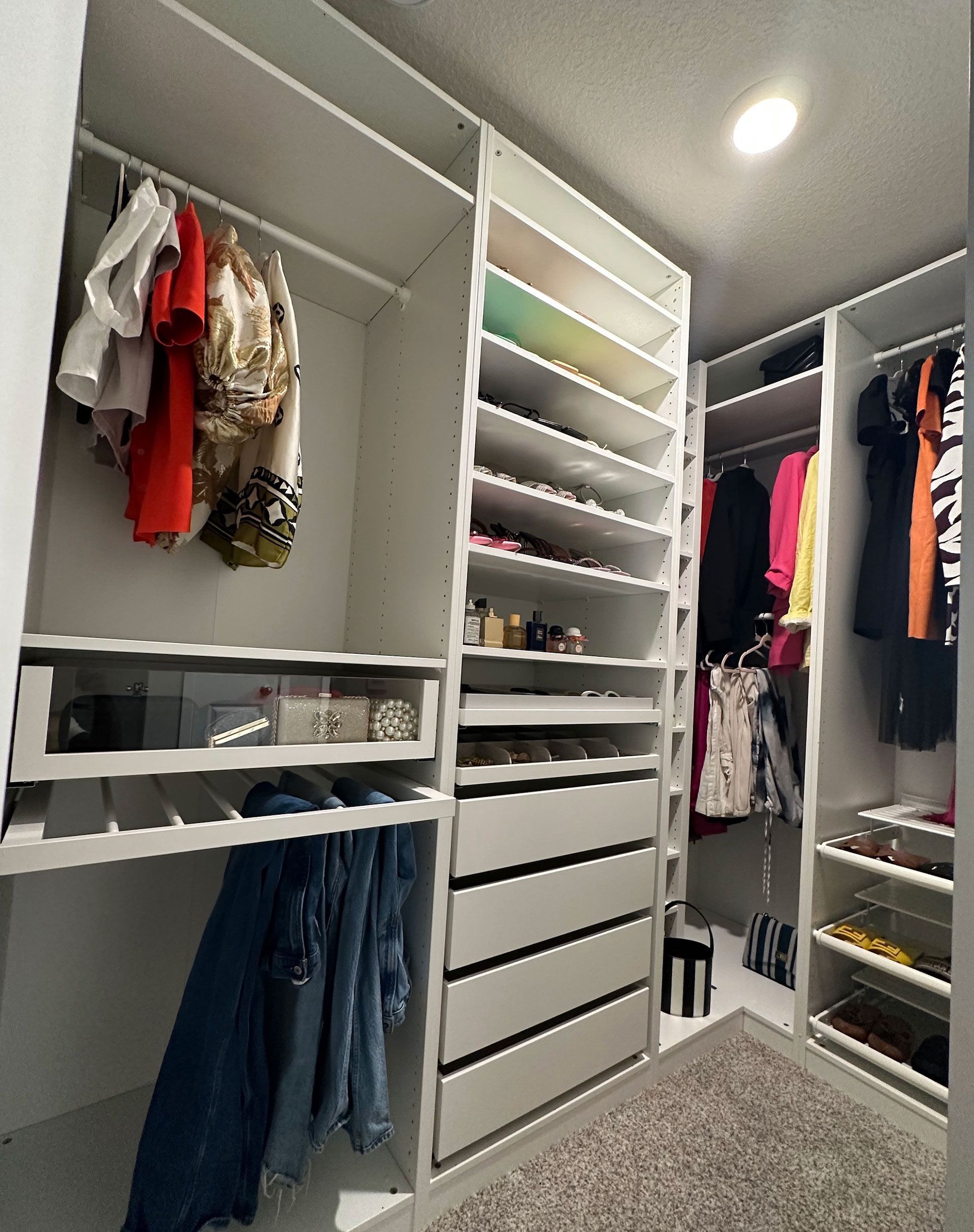 Walk-in closet with white shelves and drawers; clothes hang on rods; shoes displayed on shelves; neutral carpet.