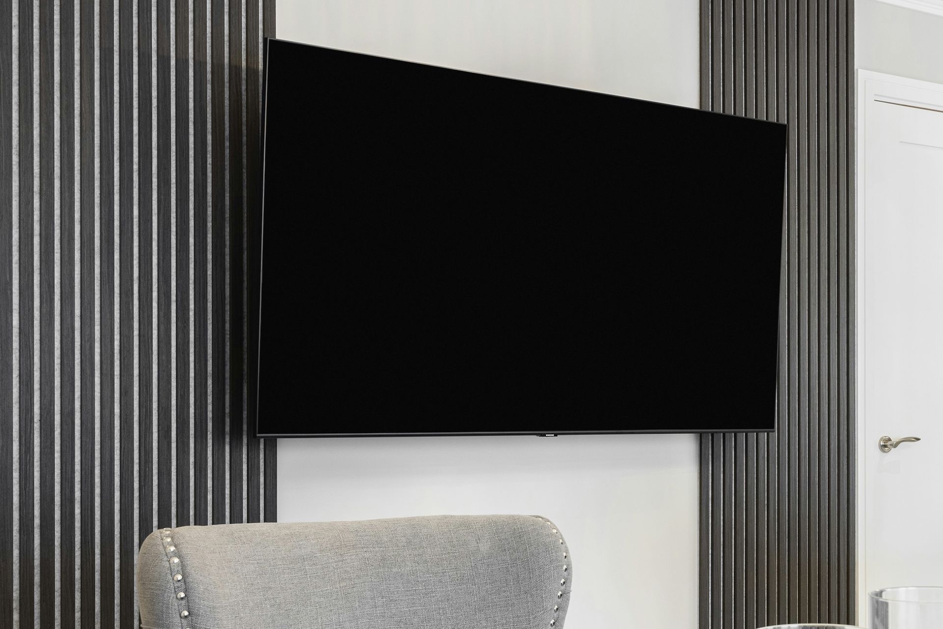 Large black TV mounted on a wall with gray vertical panels, above a gray chair.