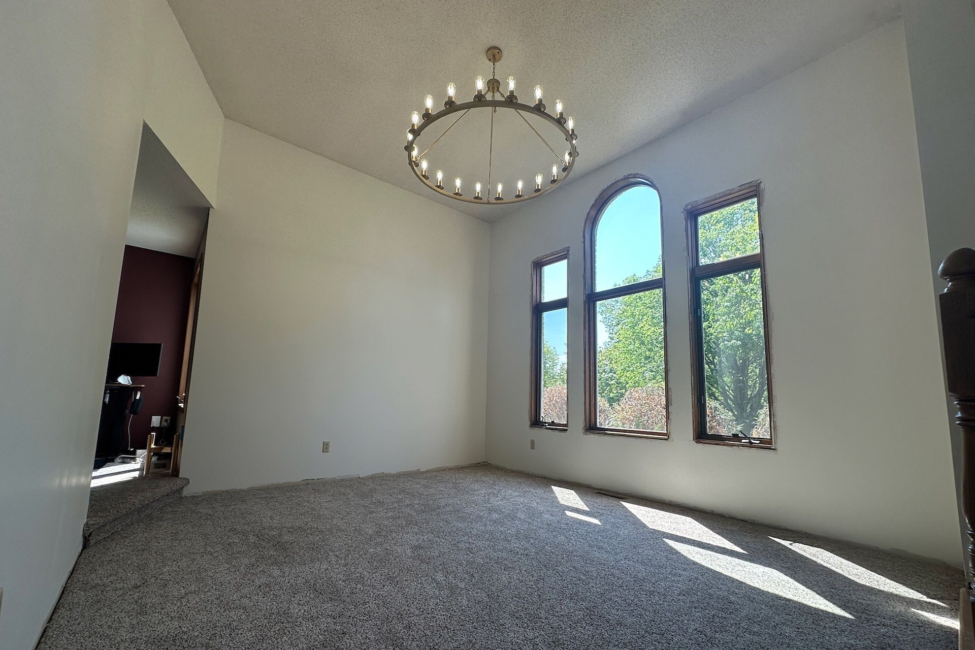 Empty room with high ceilings, arched window, chandelier, and gray carpet.