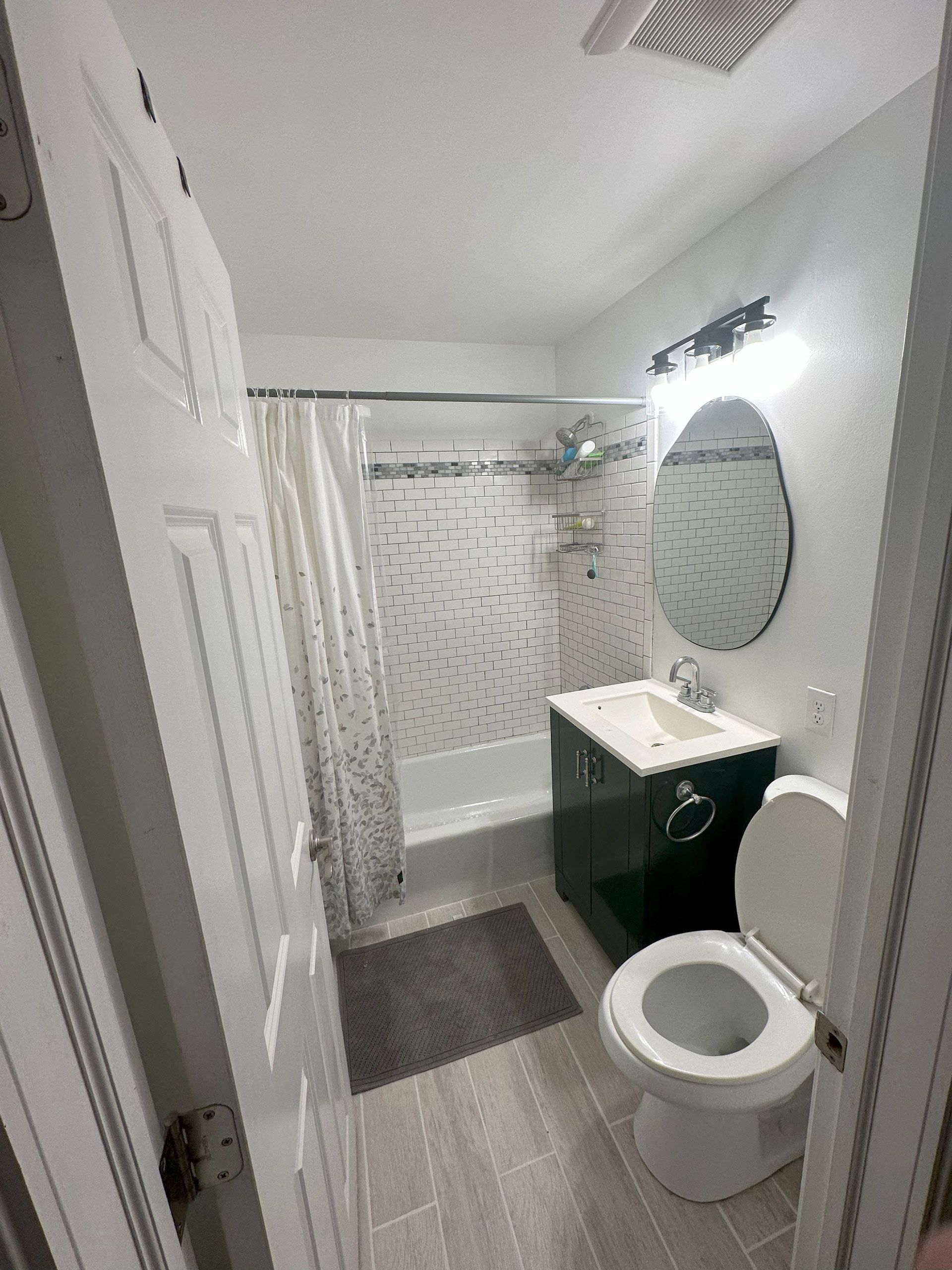 Bathroom with white and gray tiled walls, vanity, toilet, shower, and oval mirror.