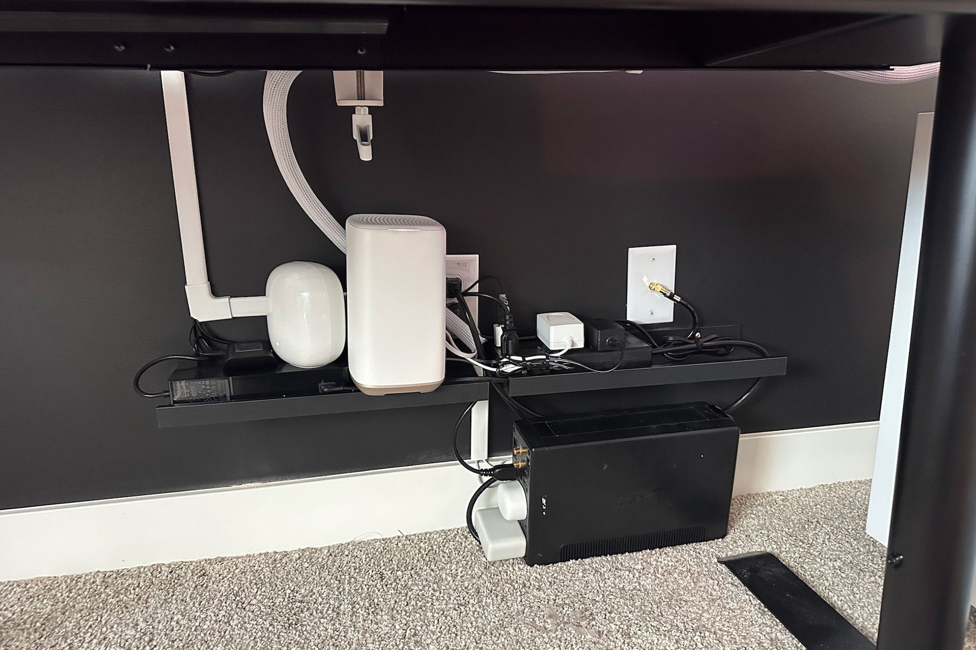 Under-desk setup with router, power strip, and other electronics on a black shelf, wires managed.