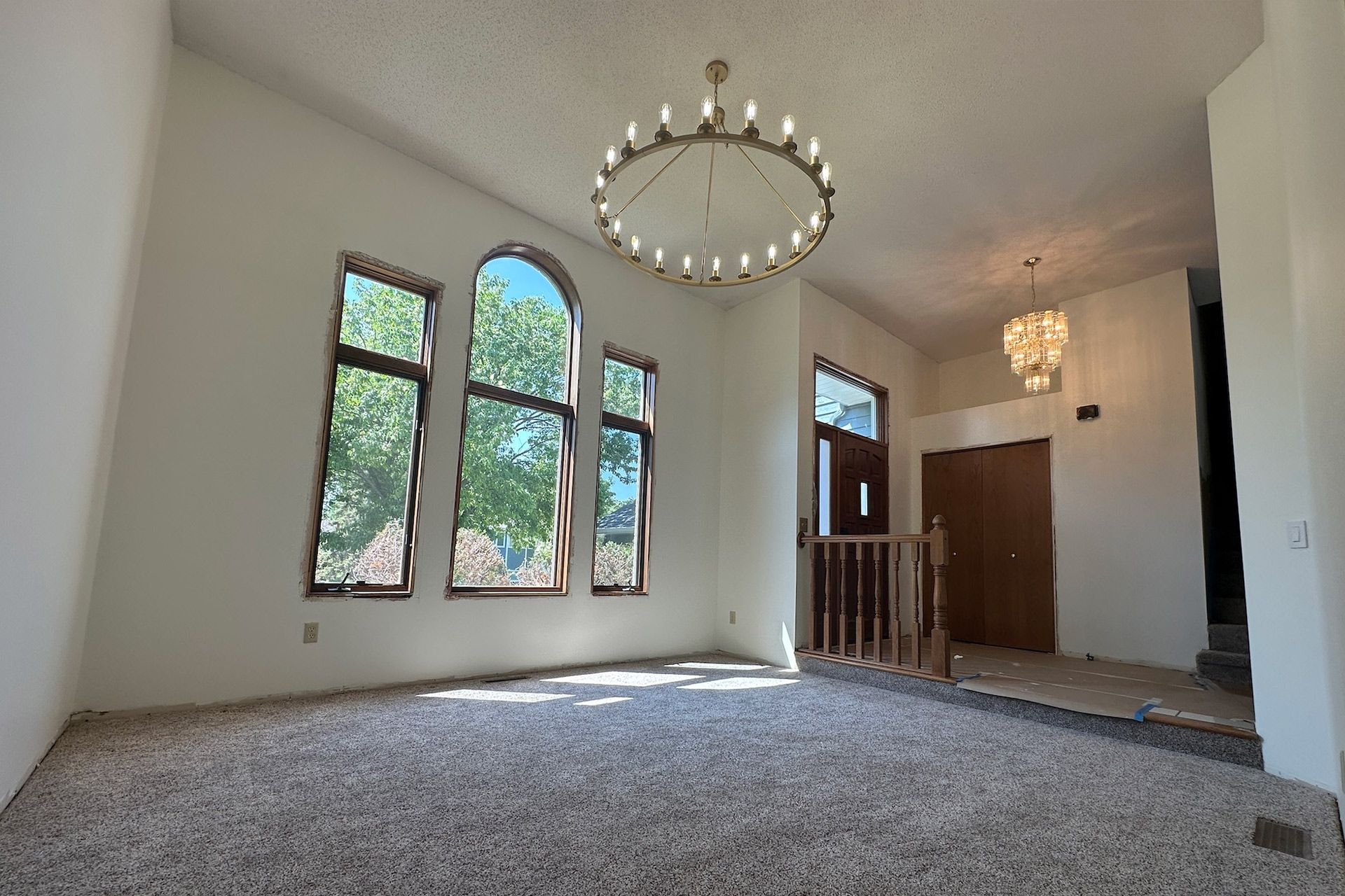 Spacious, empty room with large windows, chandeliers, and carpet flooring.
