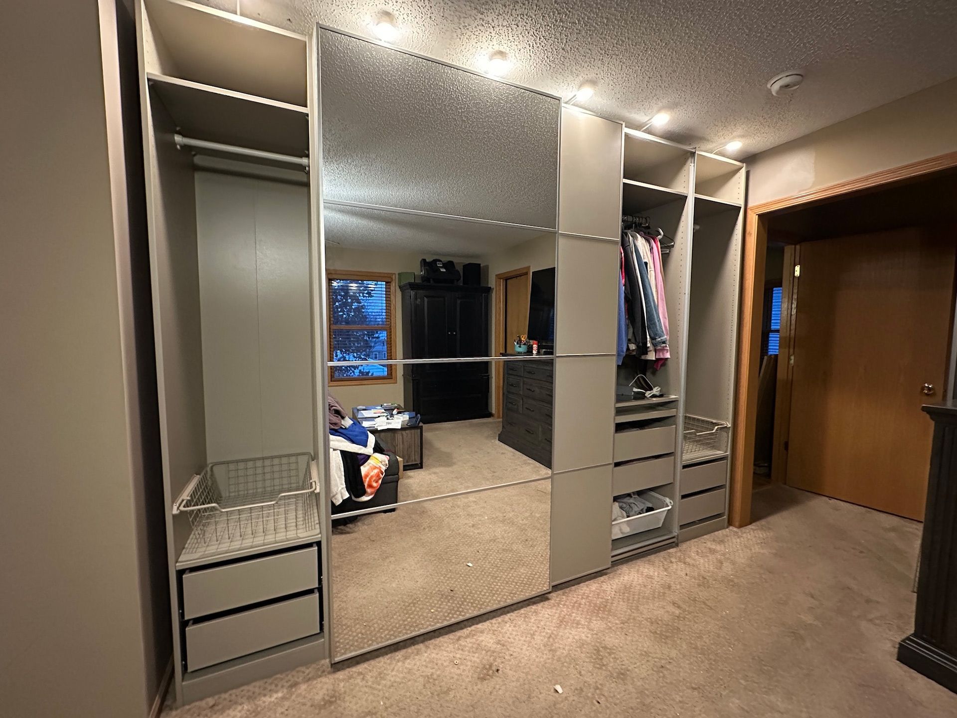 Built-in closet with mirrored sliding doors, grey shelves, drawers, and hanging rods. Lit by ceiling lights, in a bedroom.