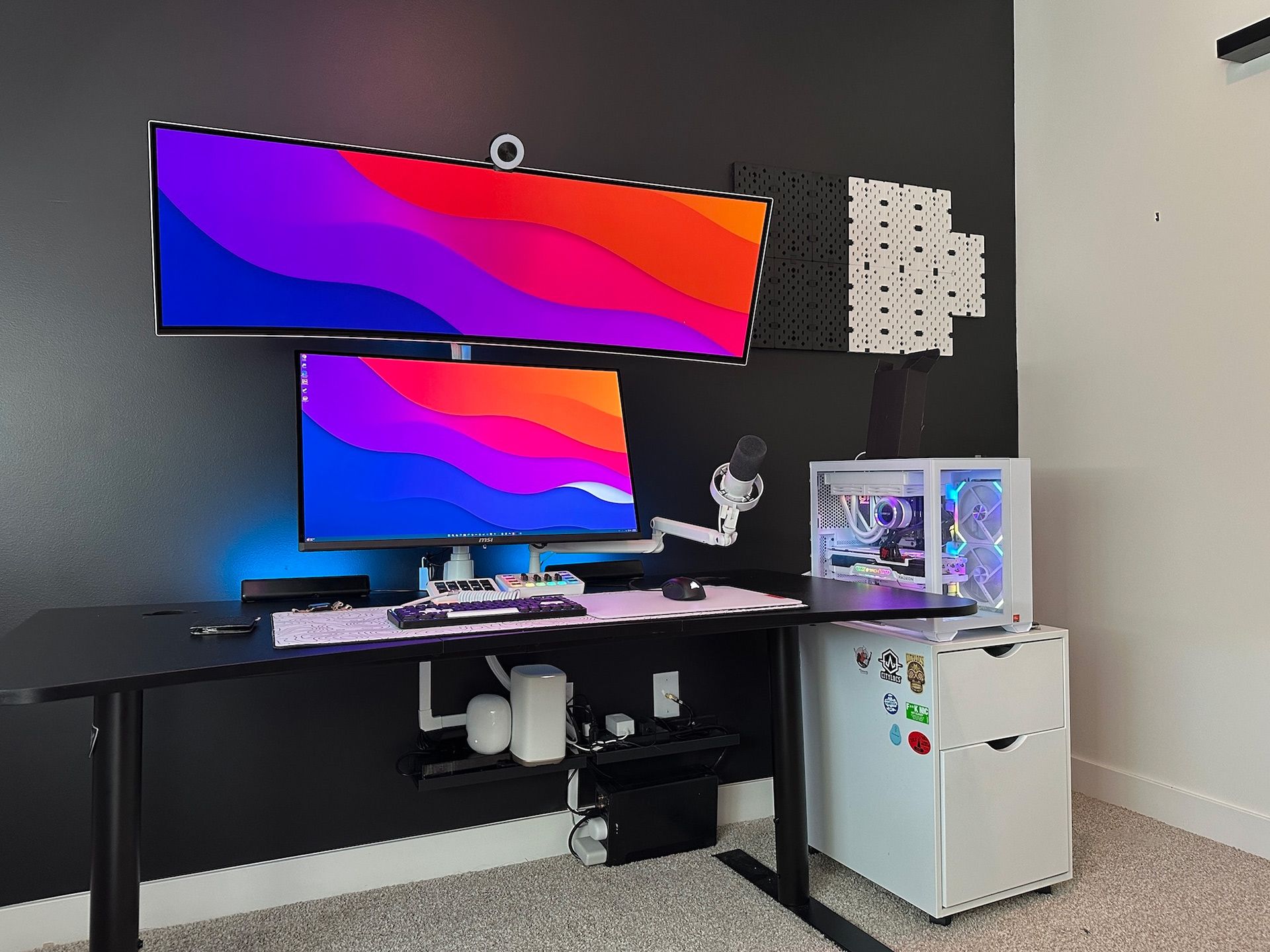 Modern desk setup with two monitors, PC, microphone, and a white filing cabinet against a black wall.