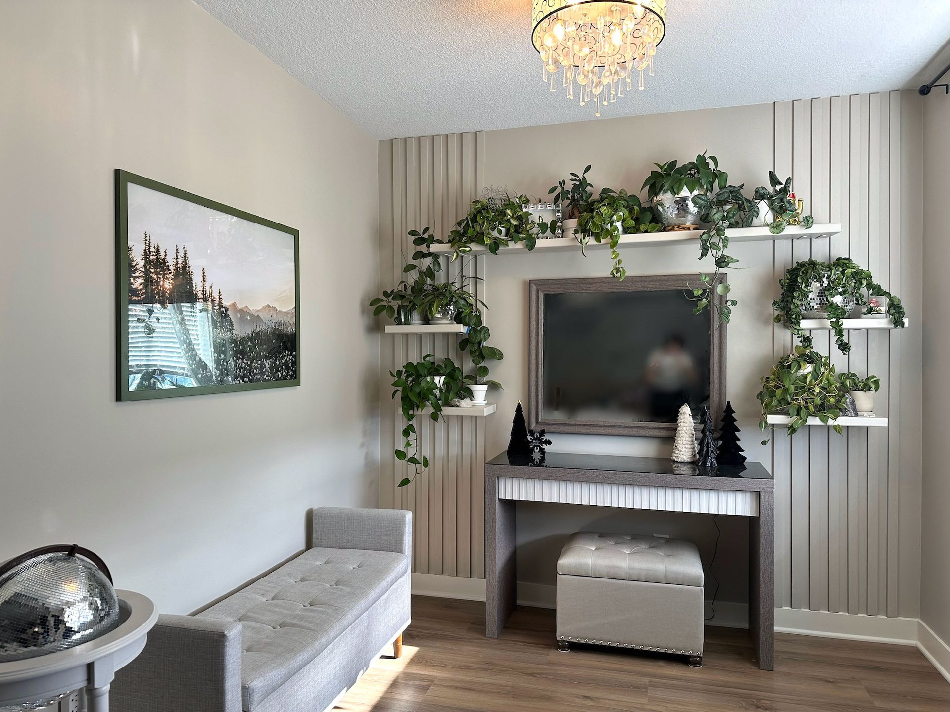 Room with plants on shelves surrounding a mirror, bench, and table. Beige walls, wood floor, and chandelier.