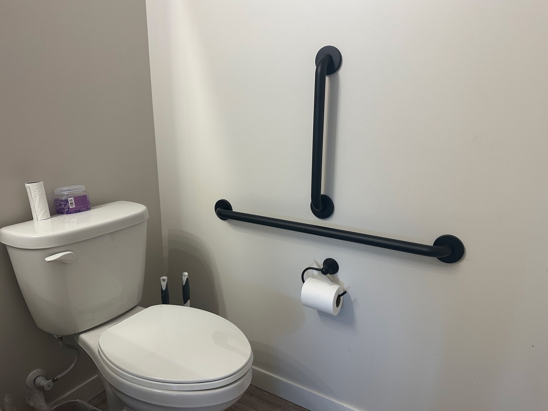 Bathroom with a toilet and black grab bars for accessibility. White toilet paper roll and gray walls.