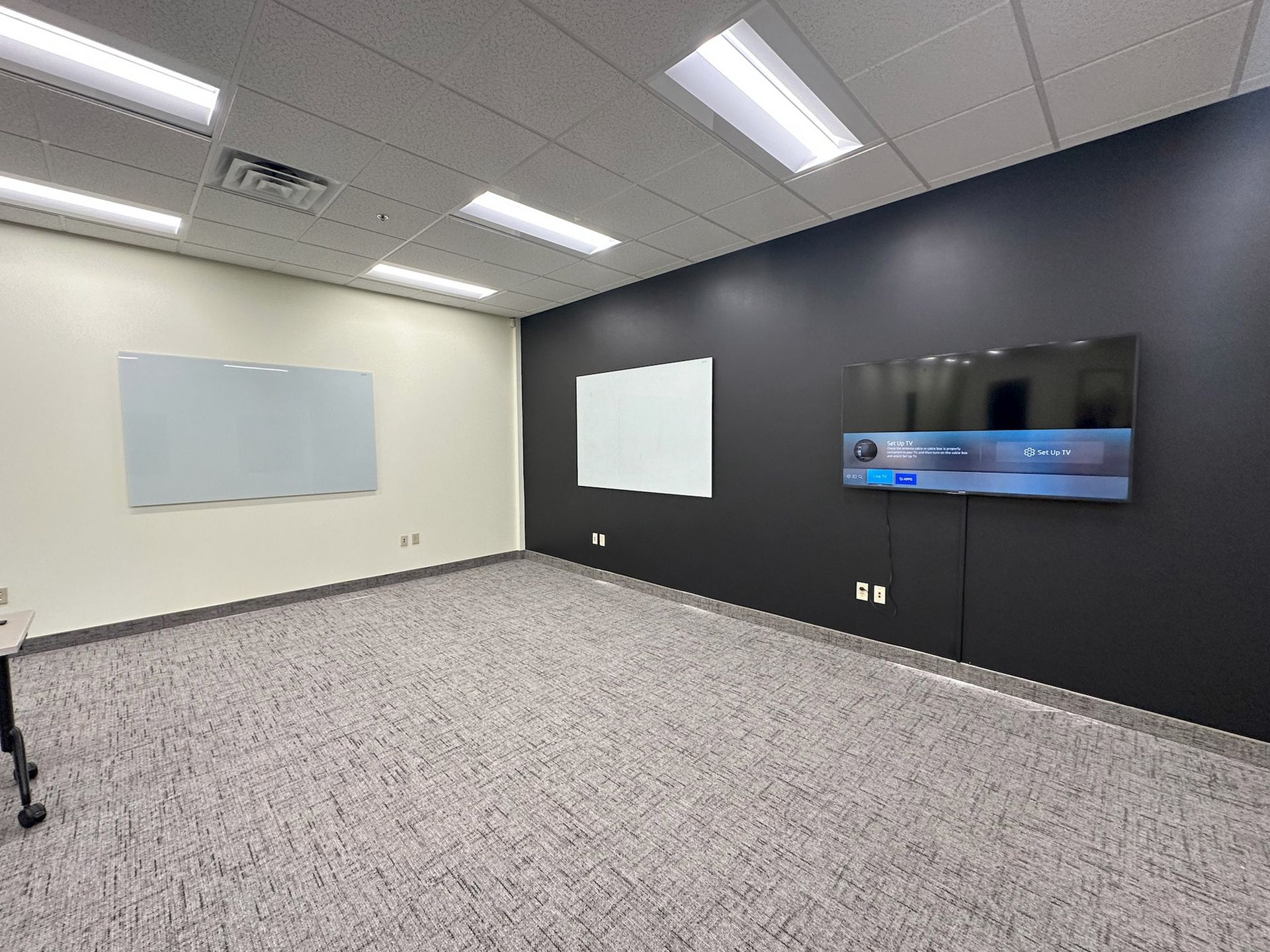 Empty office with gray carpet, white and black walls, whiteboards, and a large display screen.