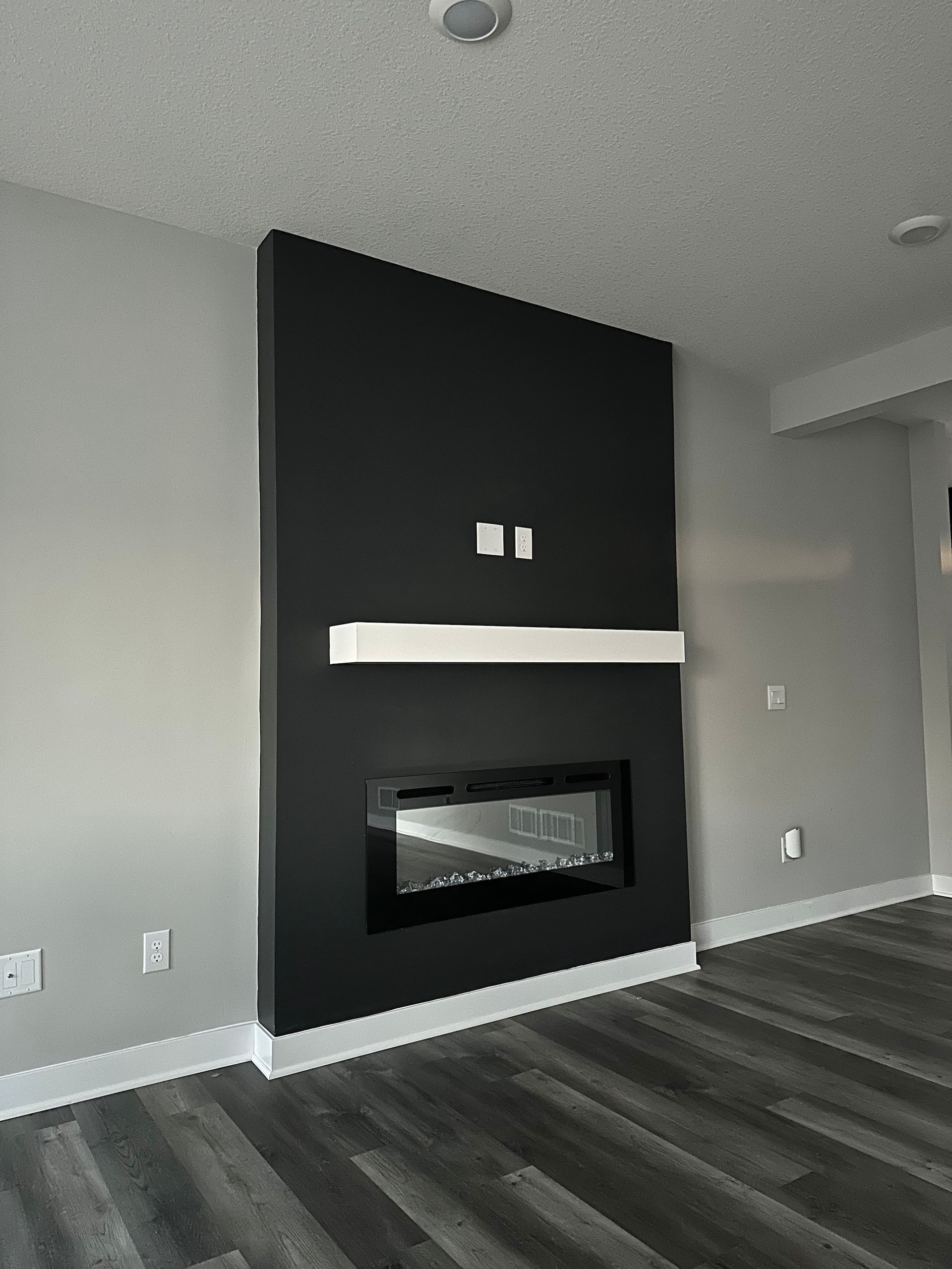 Modern living room with black fireplace built into a black wall; white mantel, grey walls, and dark flooring.