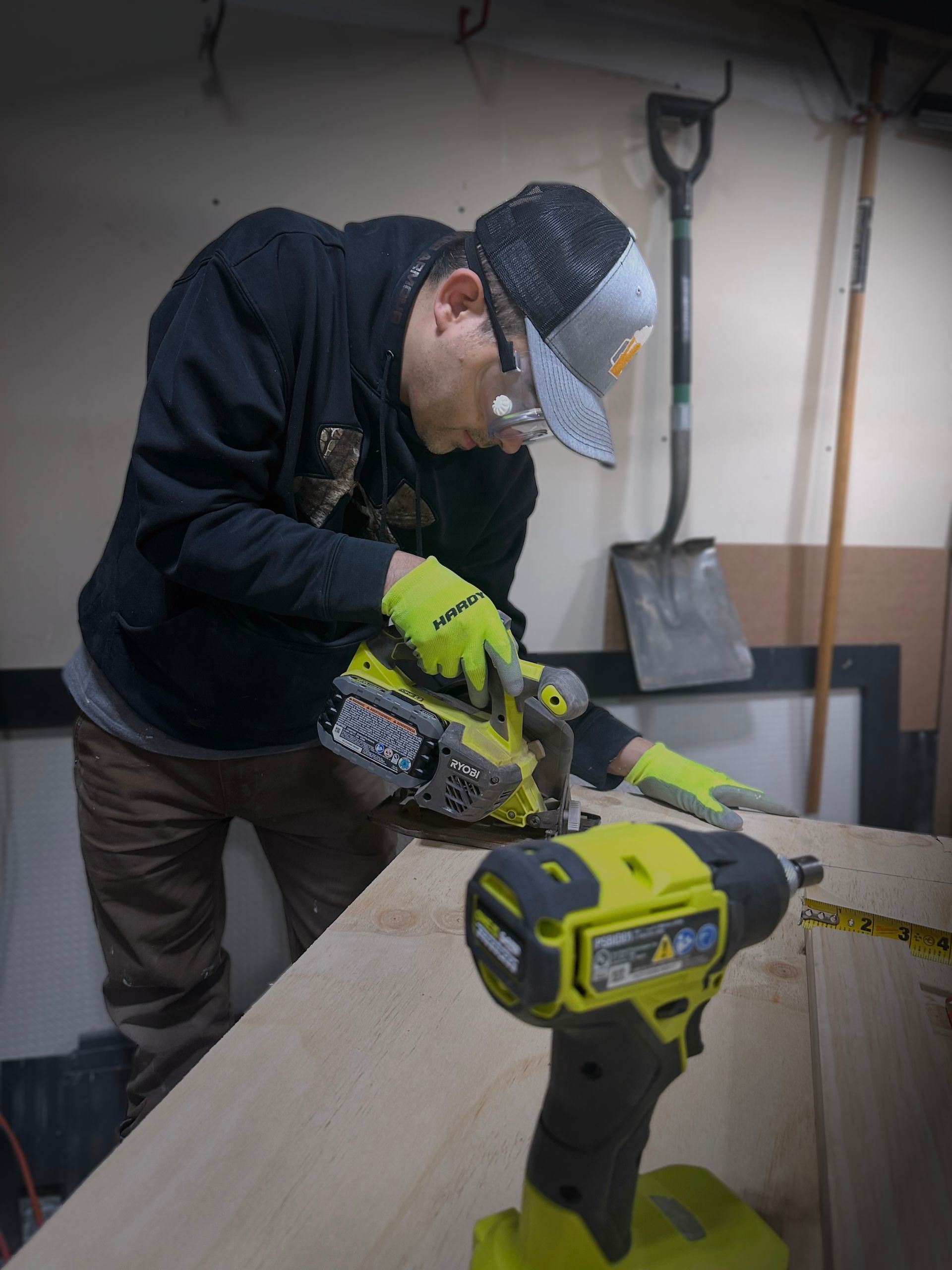 Person using a power tool to cut wood; wearing safety glasses and gloves in a workshop with a second tool in the foreground.