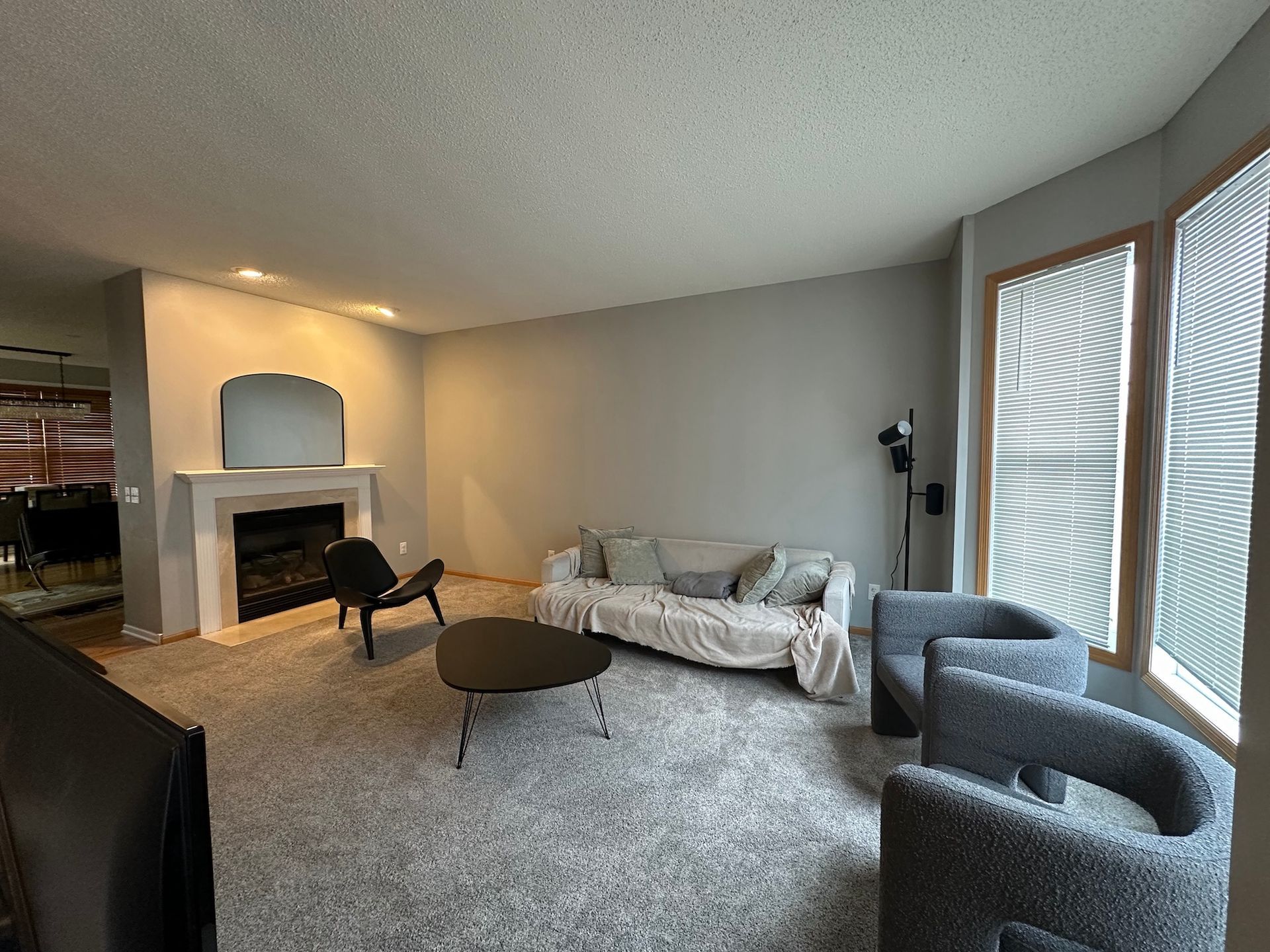 Living room with gray walls, sofa, chairs, fireplace, and large windows with blinds.