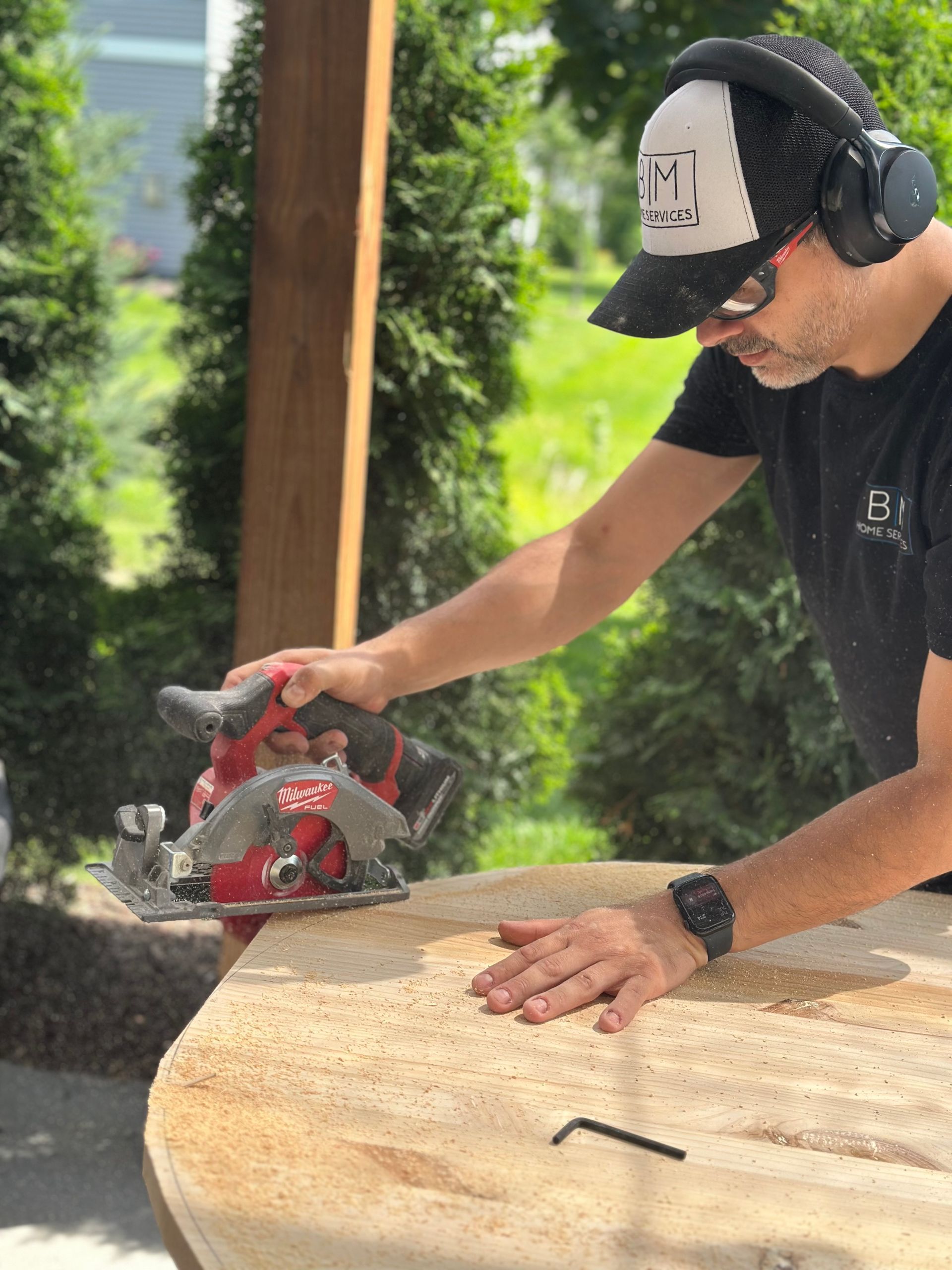 Man using a circular saw on a wooden surface outdoors, wearing hearing protection and a baseball cap.