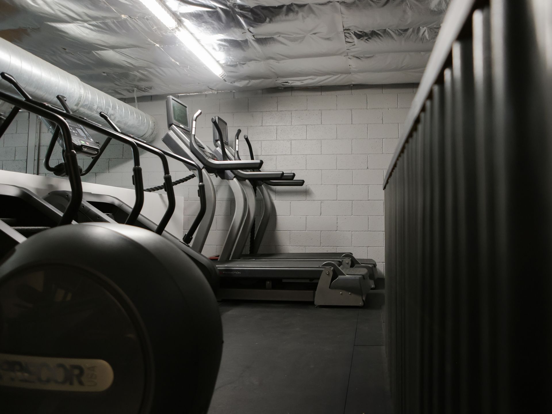 Woman swings kettlebell in a gym, others follow, performing a workout. Gym equipment in a fitness room; elliptical machine in foreground, treadmills and white brick wall in background.