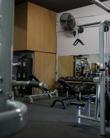 Man running on treadmill in a gym, woman behind him. Dark room, neutral expressions, exercising. Gym interior with weight machines, benches, and dumbbell racks. Dark flooring, light walls, and a large mirror.