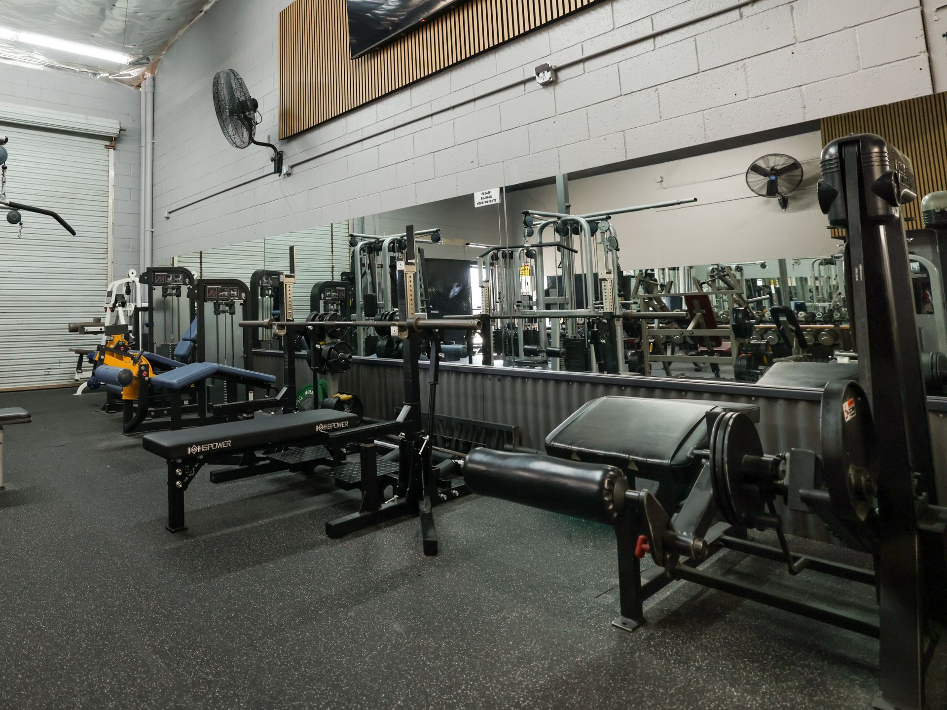 Gym interior with weight machines, benches, and a large mirror. Black equipment against gray walls and flooring. Gym interior with weight machines, benches, and a large mirror. Black equipment against gray walls and flooring.