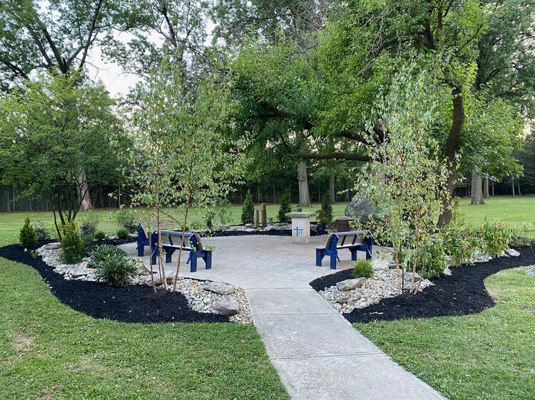 A concrete walkway leading to a picnic area in a park surrounded by trees and bushes.