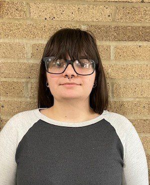 A woman wearing glasses and a nose ring is standing in front of a brick wall.