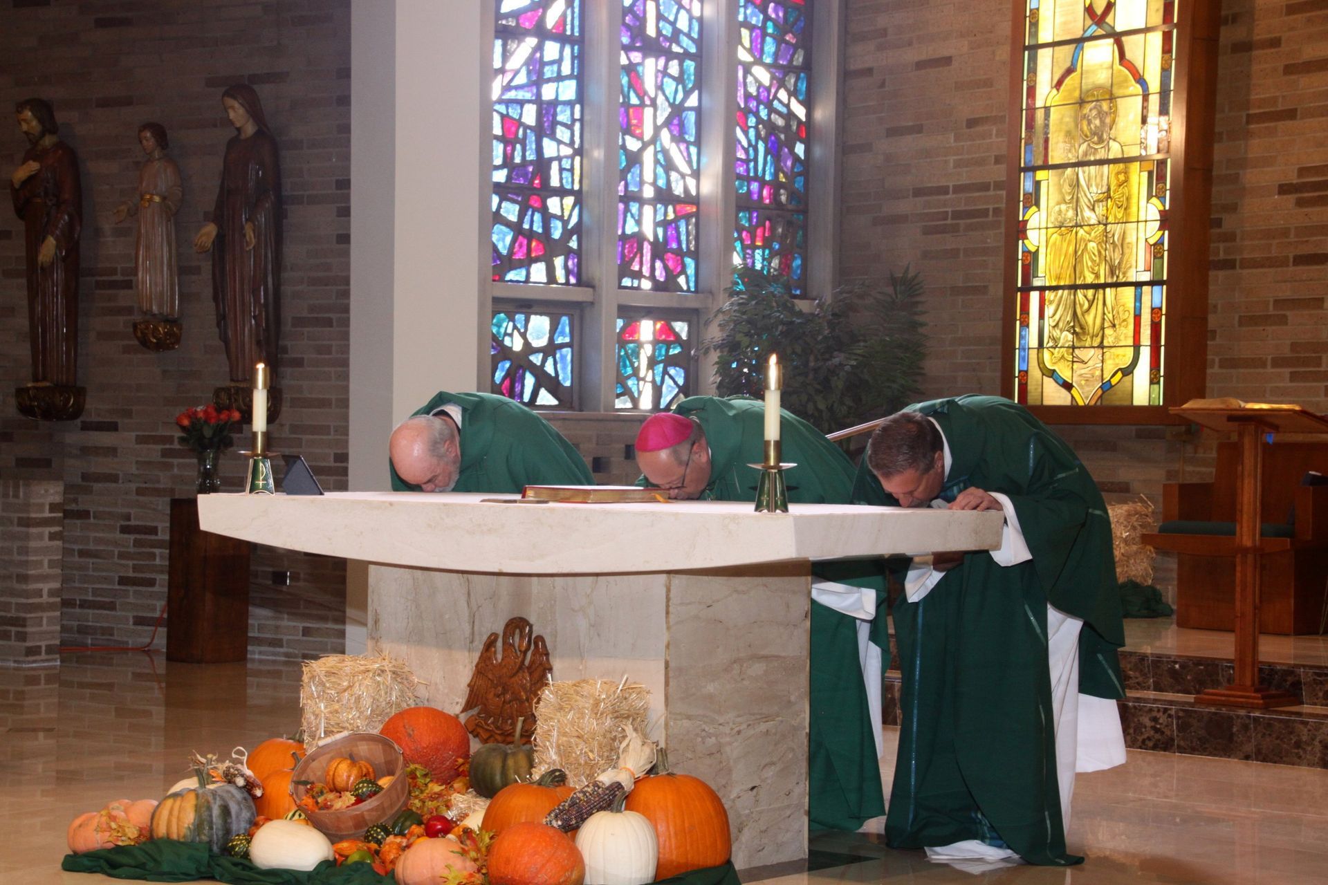 Three priests are kneeling at an altar in front of a stained-glass window.