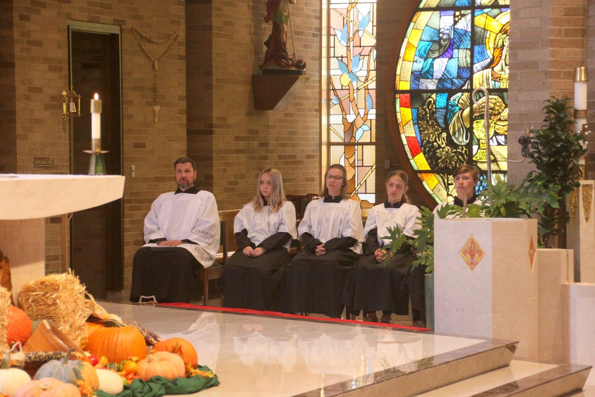 A group of altar attendants are sitting in front of a stained-glass window in a church.