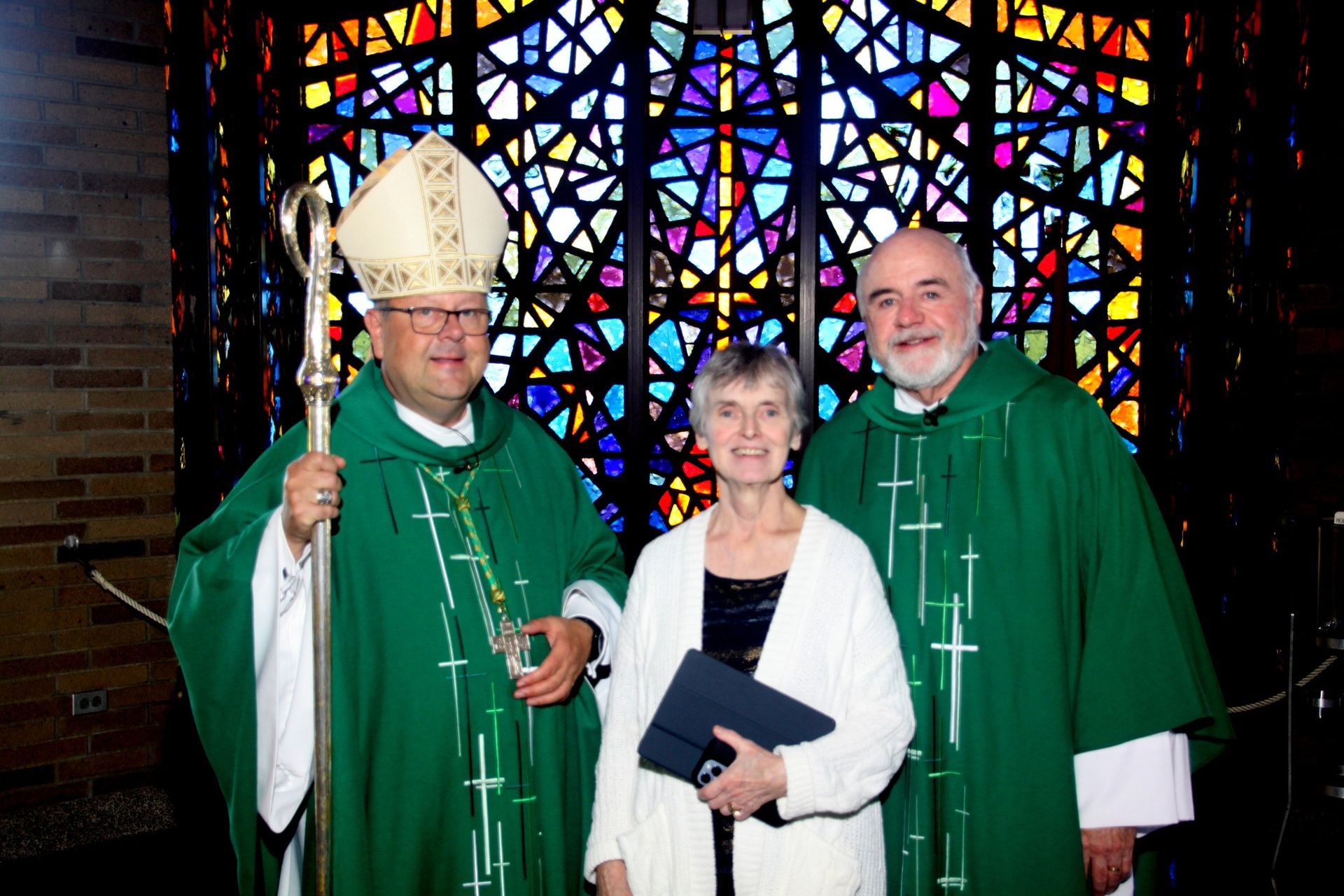 Two clergy and a woman are posing for a picture in front of a stained-glass window.