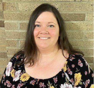 A woman in a floral shirt is smiling in front of a brick wall.