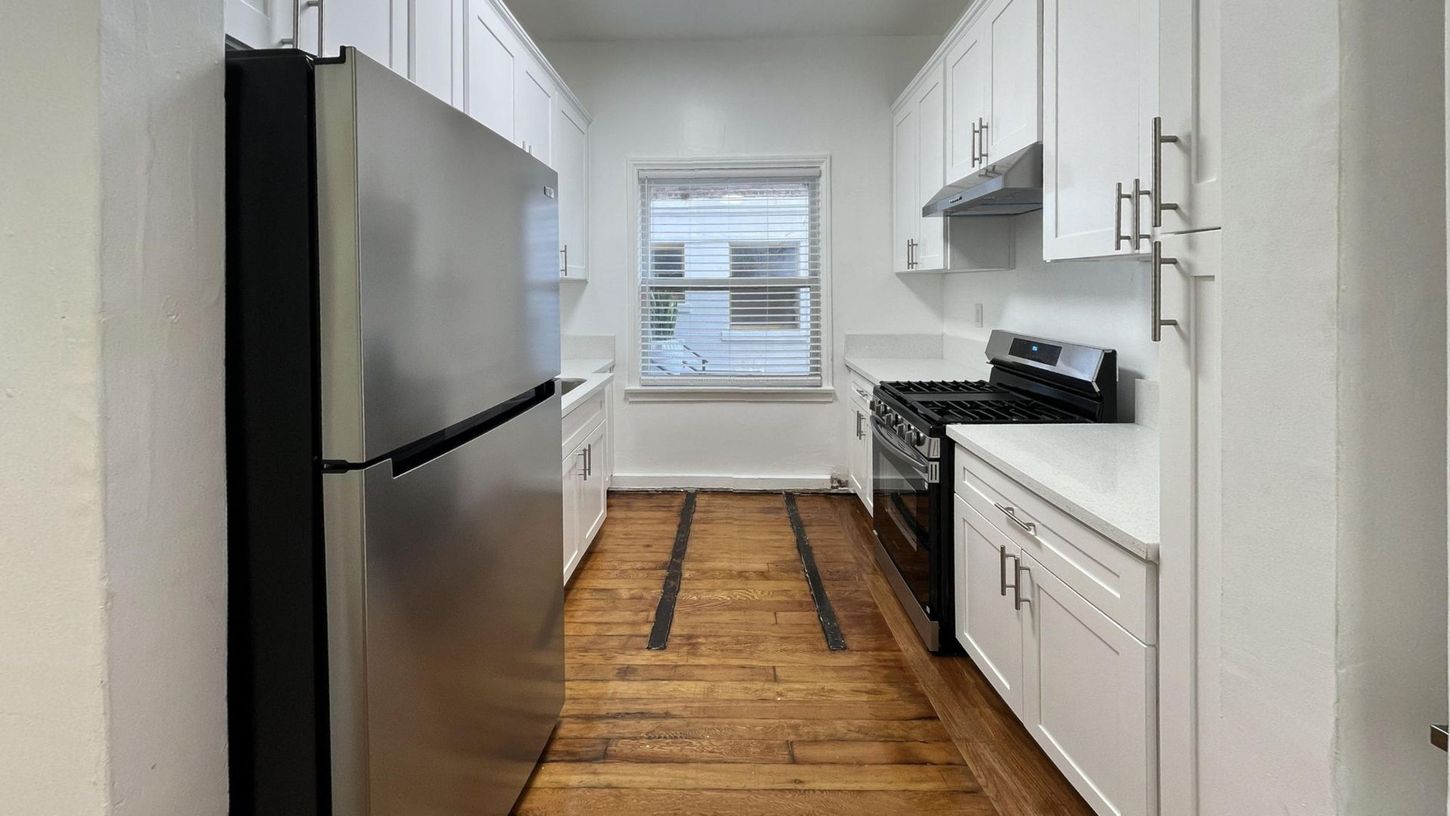 A galley kitchen with white cabinets, stainless steel refrigerator, and wood floor.