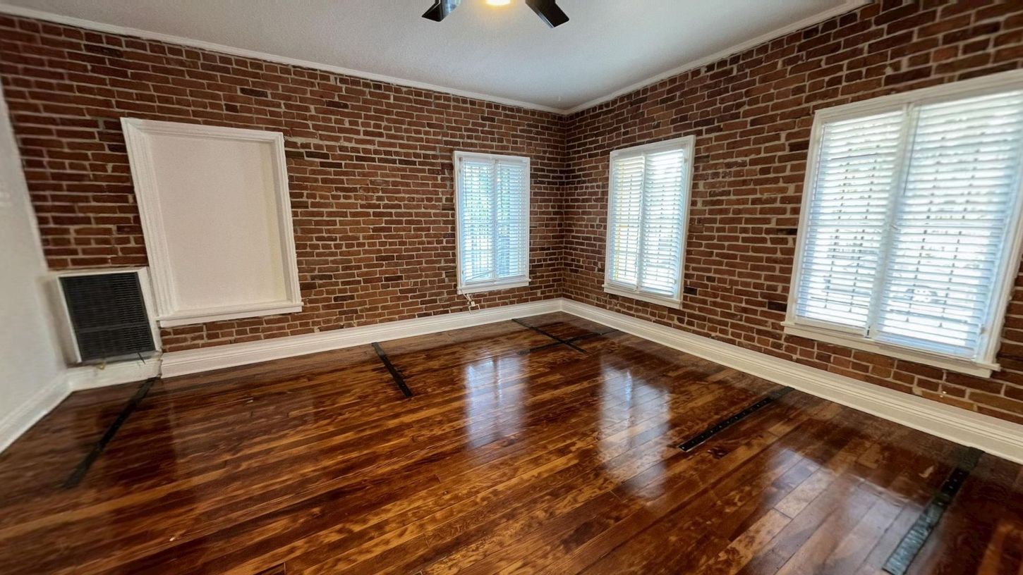 Empty room with exposed brick walls, wood floor, and three windows.